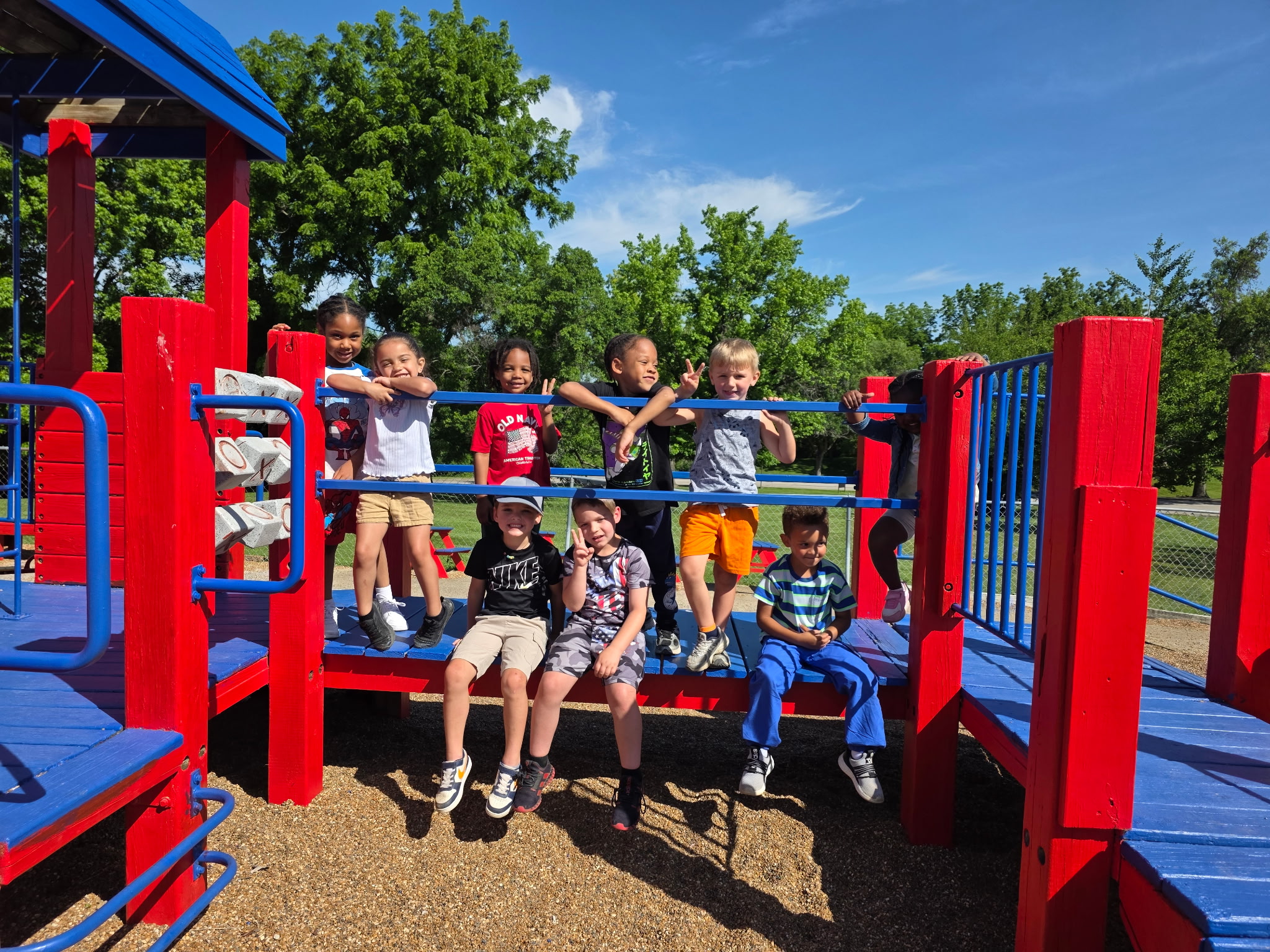Children from Little Explorers sitting and standing on the wooden playground structure outside, with green trees and blue sky behind them