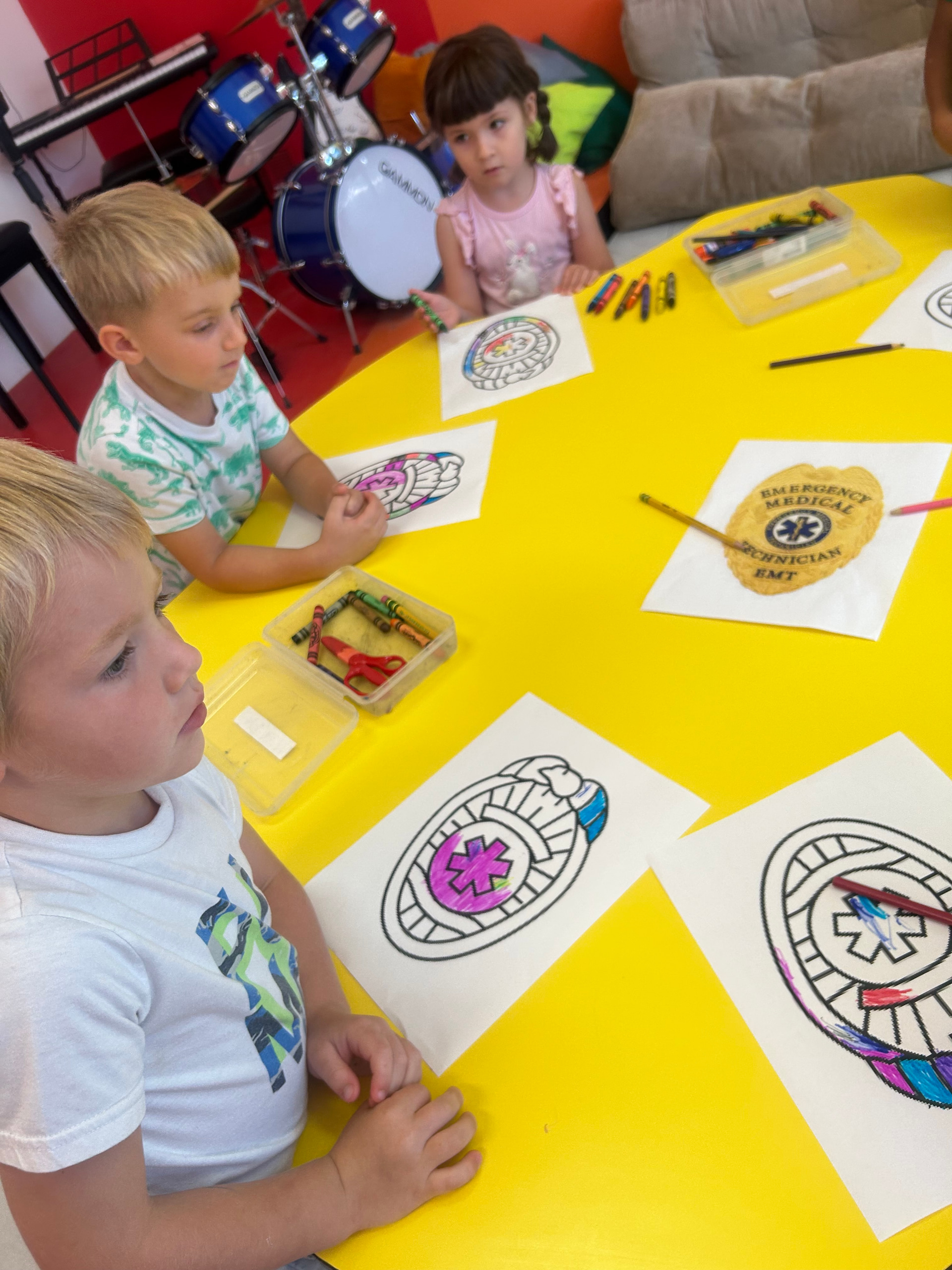 Three children coloring line drawings with crayons around a bright yellow table in a classroom with a red wall