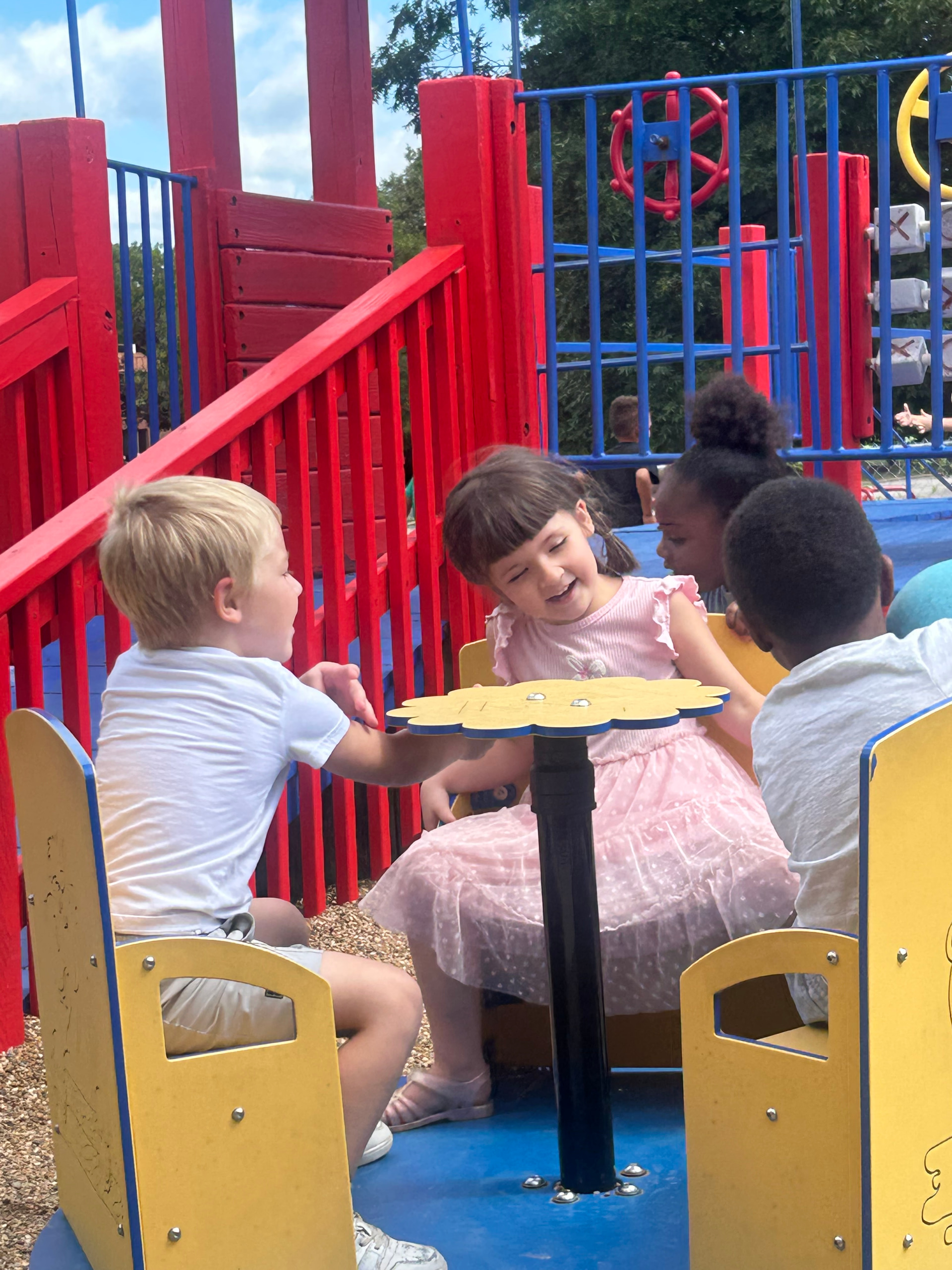 Four children from Little Explorers sitting around the yellow flower-shaped table on the playground structure, with green trees behind them