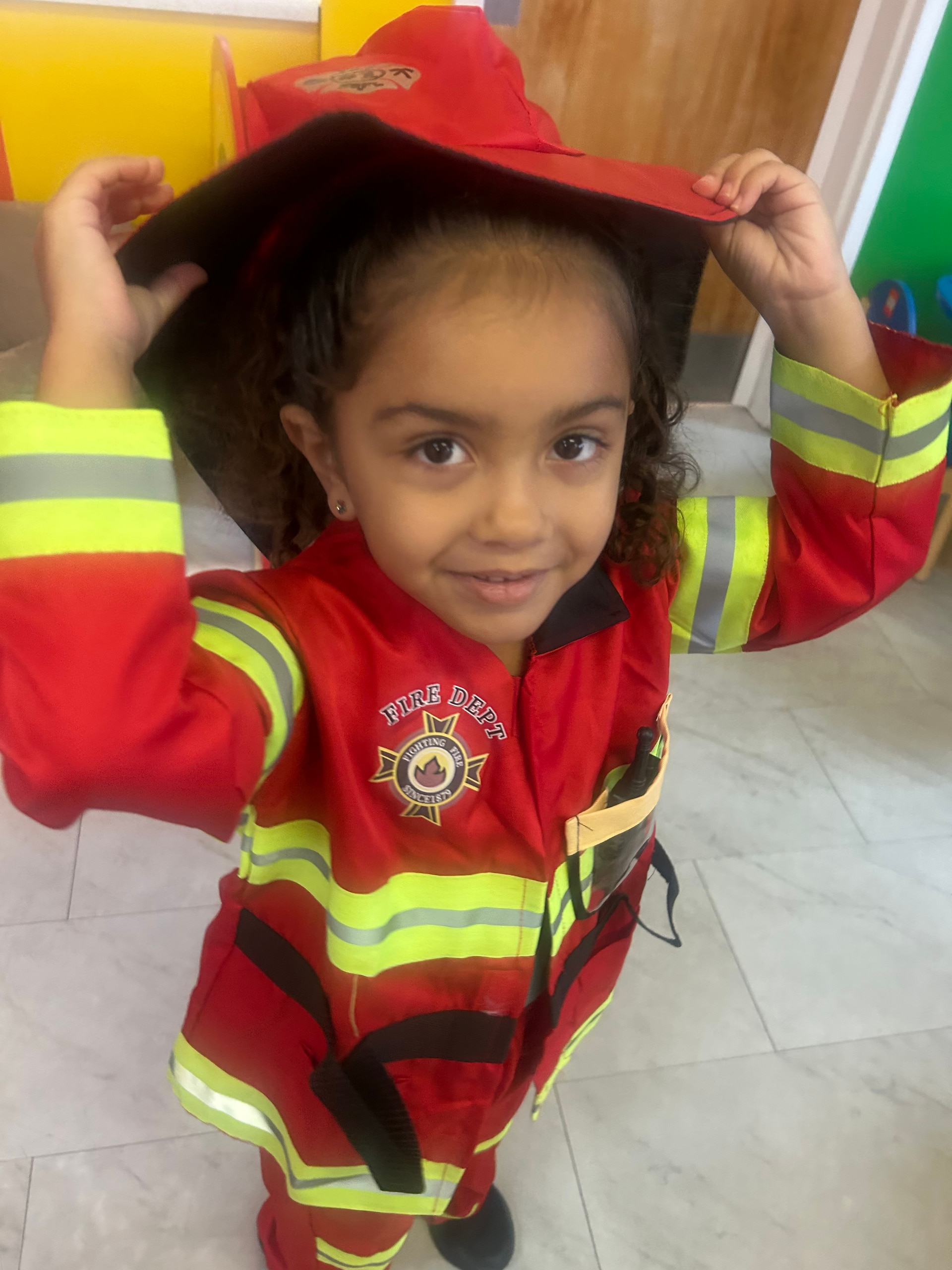 A young girl with curly hair in a red and yellow firefighter costume, holding the brim of her helmet